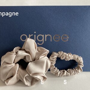 May include: Two champagne-colored satin hair scrunchies in large and small sizes. The large scrunchie is labeled L and the small one is labeled S. They are positioned in front of a dark blue card with the text champagne and the brand name orignee.