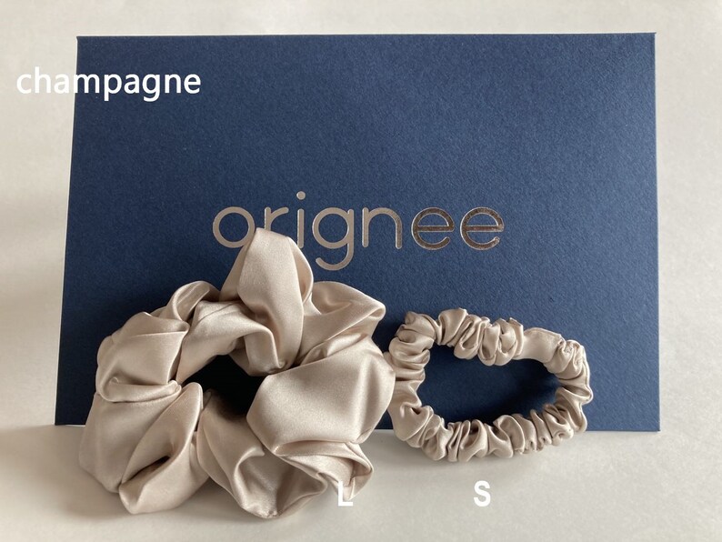 May include: Two champagne-colored satin hair scrunchies in large and small sizes. The large scrunchie is labeled L and the small one is labeled S. They are positioned in front of a dark blue card with the text champagne and the brand name orignee.