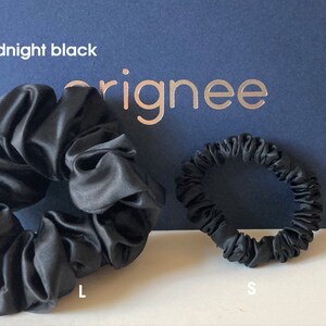 May include: Two midnight black hair scrunchies in different sizes, labeled L and S, positioned in front of a dark blue card with the text midnight black and orignee printed on it. The scrunchies have a smooth, satin-like finish and gathered fabric.