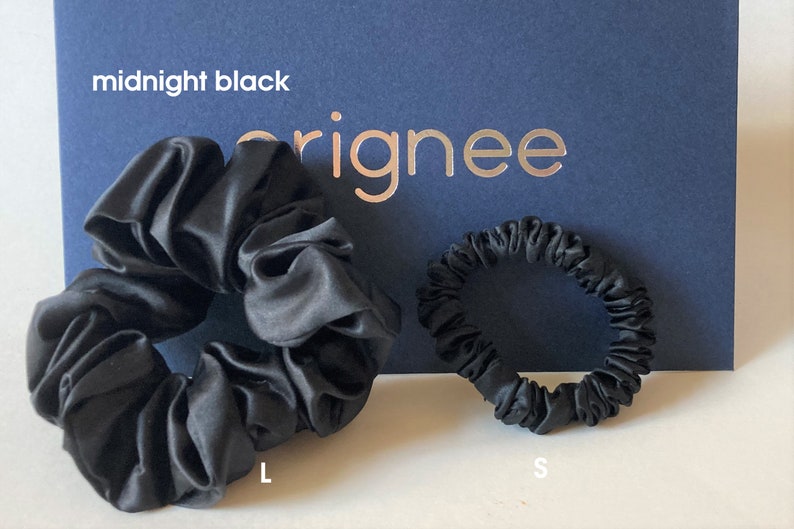 May include: Two midnight black hair scrunchies in different sizes, labeled L and S, positioned in front of a dark blue card with the text midnight black and orignee printed on it. The scrunchies have a smooth, satin-like finish and gathered fabric.