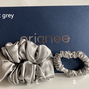 May include: Two light grey hair scrunchies in different sizes, labeled L and S, displayed against a dark blue card with the text "orignee" and "light grey" printed on it. The scrunchies have a smooth, lustrous finish and gathered fabric texture.