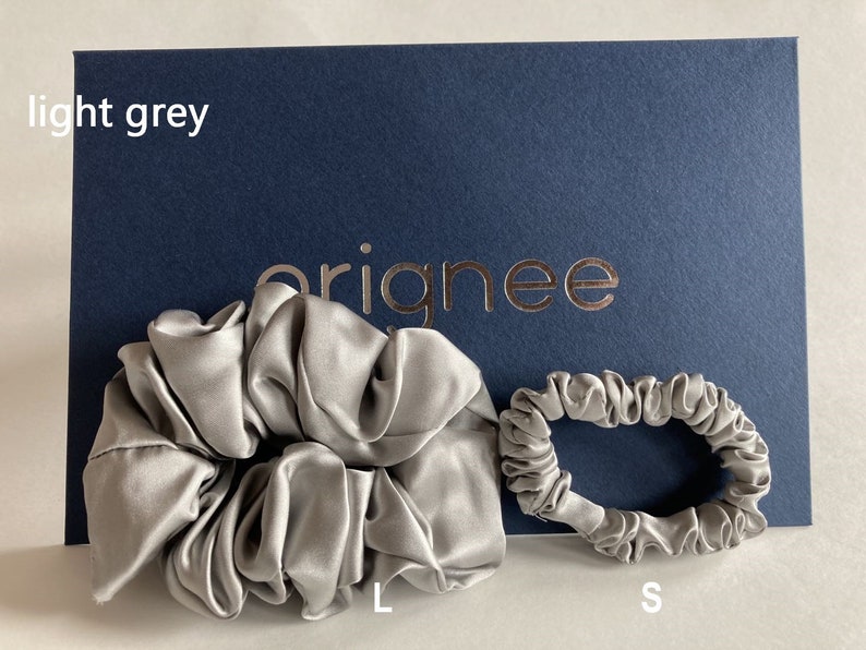 May include: Two light grey hair scrunchies in different sizes, labeled L and S, displayed against a dark blue card with the text "orignee" and "light grey" printed on it. The scrunchies have a smooth, lustrous finish and gathered fabric texture.