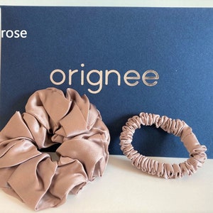 May include: Two old rose colored hair accessories, one large scrunchie and one thin hair tie, positioned in front of a dark blue rectangular card with the text originee printed in gold and the words old rose printed in white.