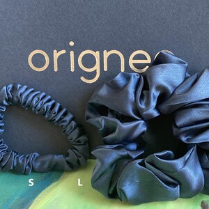 May include: Two navy blue satin hair scrunchies of different sizes, labeled S and L, positioned in front of a black card with the gold text origines. The fabric has a smooth, reflective finish with gathered folds throughout the circular bands.