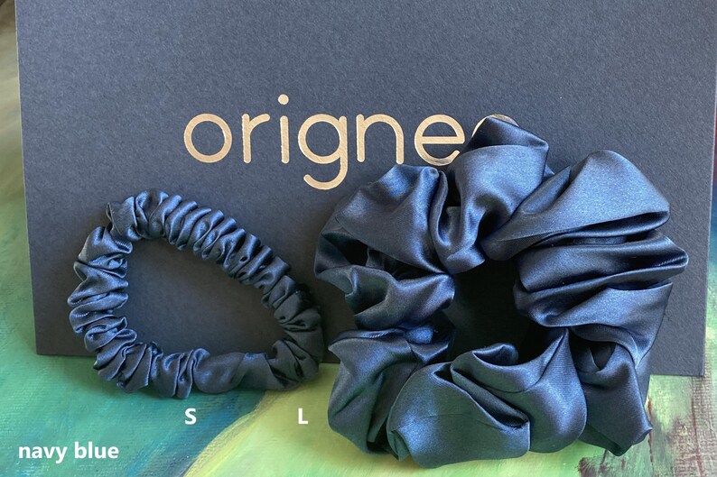 May include: Two navy blue satin hair scrunchies of different sizes, labeled S and L, positioned in front of a black card with the gold text origines. The fabric has a smooth, reflective finish with gathered folds throughout the circular bands.