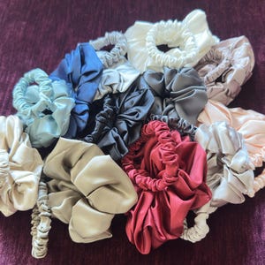May include: A collection of satin hair scrunchies in various colors including white, cream, beige, light blue, dark blue, grey, black, pink, and deep red. Each scrunchie features a gathered, pleated fabric design with an elastic interior band.