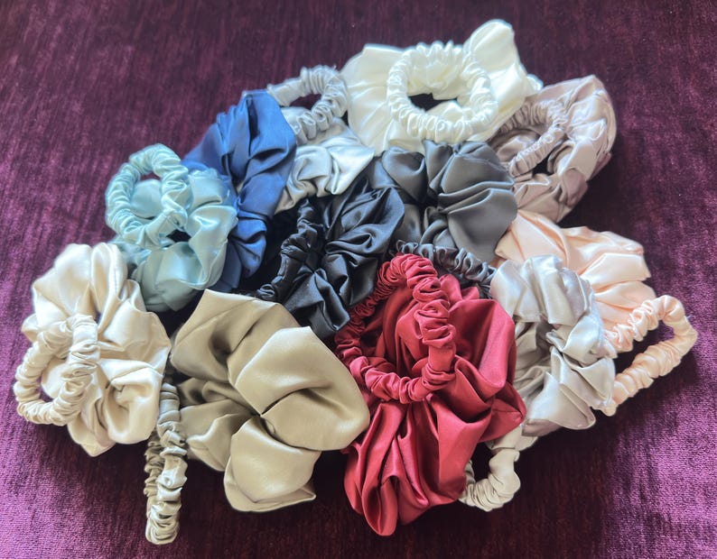 May include: A collection of satin hair scrunchies in various colors including white, cream, beige, light blue, dark blue, grey, black, pink, and deep red. Each scrunchie features a gathered, pleated fabric design with an elastic interior band.