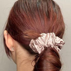 May include: A dusty rose-colored satin scrunchie with a gathered, pleated texture. The fabric has a smooth, reflective sheen and is wrapped around a ponytail of dark reddish-brown hair, securing the hair in a low, gathered style.