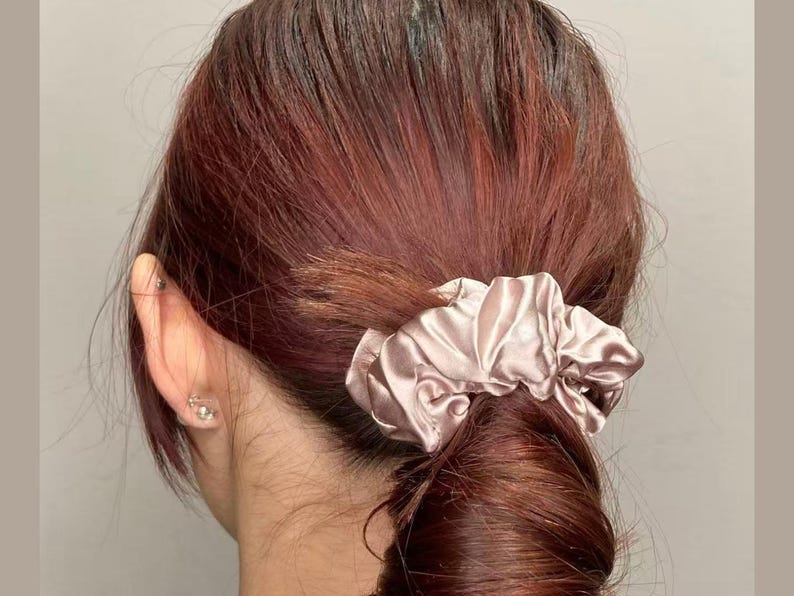 May include: A dusty rose-colored satin scrunchie with a gathered, pleated texture. The fabric has a smooth, reflective sheen and is wrapped around a ponytail of dark reddish-brown hair, securing the hair in a low, gathered style.