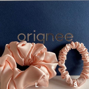 May include: Two light pink satin hair scrunchies, one large and one small, positioned in front of a dark blue card with the word originee printed in metallic gold. The text pink is visible in the top left, with L and S labels below.