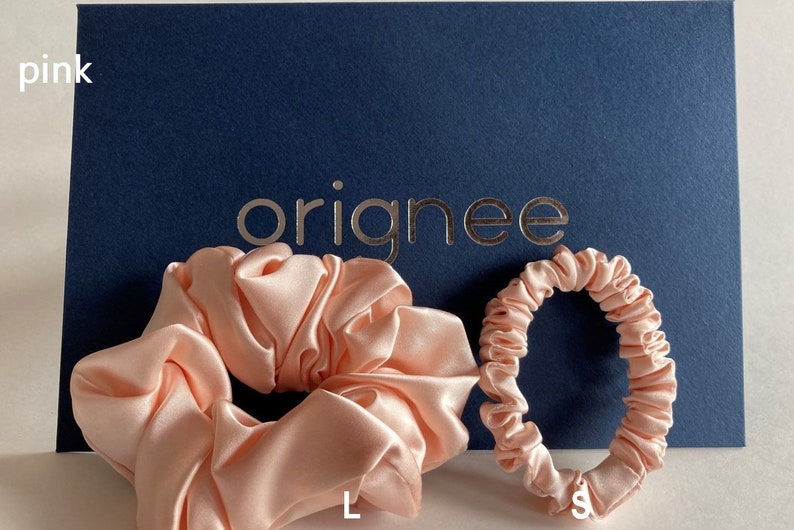 May include: Two light pink satin hair scrunchies, one large and one small, positioned in front of a dark blue card with the word originee printed in metallic gold. The text pink is visible in the top left, with L and S labels below.