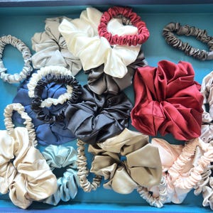 May include: A collection of various fabric hair scrunchies in a blue box. The assortment includes satin-like textures in colors such as cream, deep red, navy blue, charcoal gray, light blue, beige, and pale pink, featuring both thin and wide styles.