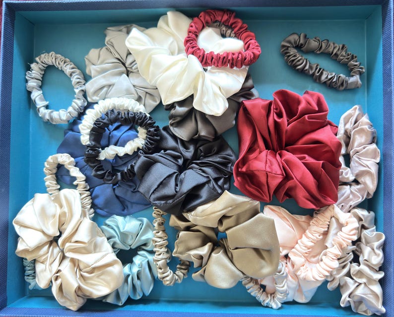 May include: A collection of various fabric hair scrunchies in a blue box. The assortment includes satin-like textures in colors such as cream, deep red, navy blue, charcoal gray, light blue, beige, and pale pink, featuring both thin and wide styles.