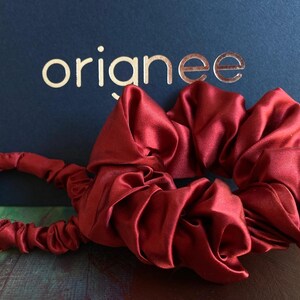 May include: A deep red satin scrunchie with a gathered, elasticized fabric design. The accessory features a smooth, reflective texture and is positioned in front of a dark blue background with the gold-colored text orianee printed on it.