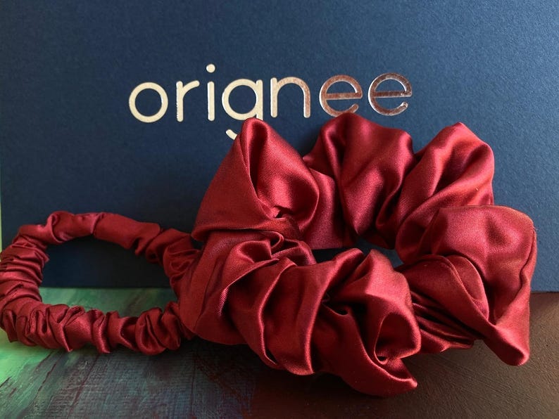 May include: A deep red satin scrunchie with a gathered, elasticized fabric design. The accessory features a smooth, reflective texture and is positioned in front of a dark blue background with the gold-colored text orianee printed on it.