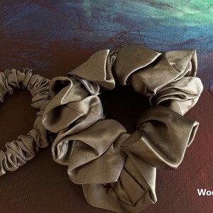 May include: Two wood brown hair scrunchies with a smooth, gathered fabric texture. One scrunchie is smaller, labeled with the letter S, and the other is larger. The items are displayed against a dark, abstract painted background.
