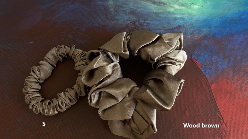 May include: Two wood brown hair scrunchies with a smooth, gathered fabric texture. One scrunchie is smaller, labeled with the letter S, and the other is larger. The items are displayed against a dark, abstract painted background.