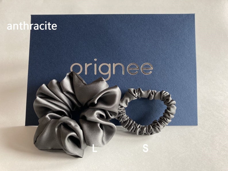 May include: A large and a small anthracite-colored satin hair scrunchie set. The items are displayed in front of a dark blue card featuring the text "orignee" in gold lettering, with the word "anthracite" printed in white in the top left corner.