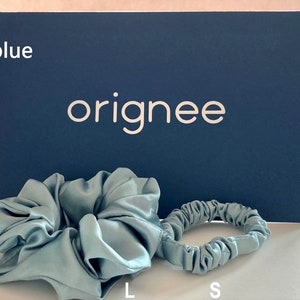 May include: Two light blue hair scrunchies, one large and one small, positioned in front of a dark blue rectangular card with the white text originee and the words light blue, L, and S printed on the card.