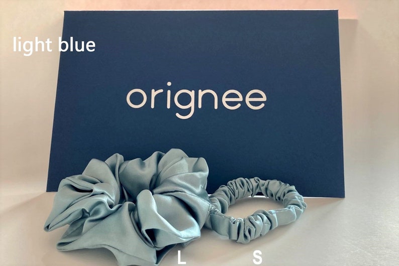 May include: Two light blue hair scrunchies, one large and one small, positioned in front of a dark blue rectangular card with the white text originee and the words light blue, L, and S printed on the card.