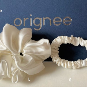 May include: Two white satin hair scrunchies, one large and one small, positioned in front of a dark blue card with the text "originee" in silver lettering. The large scrunchie is voluminous and gathered, while the small scrunchie is thinner.