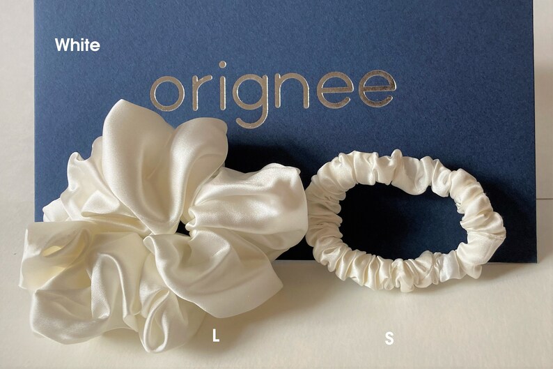 May include: Two white satin hair scrunchies, one large and one small, positioned in front of a dark blue card with the text "originee" in silver lettering. The large scrunchie is voluminous and gathered, while the small scrunchie is thinner.