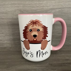 May include: White ceramic mug with a pink rim and handle. A cartoon brown dog with brown eyes peeks over the rim of the mug. The text "Jojo's Mom" is printed on the mug.