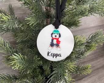 May include: White ceramic Christmas ornament with a cartoon dog wearing a green hat and red scarf. The ornament has the name "Keppa" written on it.