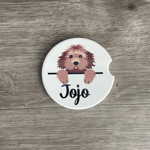 May include: White ceramic coaster with a brown and white dog peeking over the top. The dog's name, "Jojo", is written in black cursive lettering.