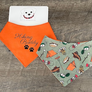 May include: Orange and green dog bandana with the text "Hiking Buddy" and paw prints. The bandana is on a wood background with a white card that says "Katie Anderson" and has a cartoon dog on it.