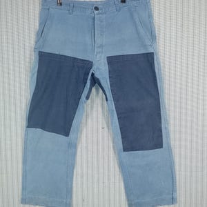 Può includere: Pantaloni di jeans azzurri con due toppe blu scuro sul davanti. I pantaloni sono appesi a una gruccia di metallo. Hanno una chiusura con bottone e cerniera e tasche.