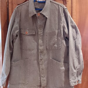 Peut inclure: Une veste de travail grise avec une fermeture boutonnée, deux poches de poitrine et deux poches inférieures. La veste est suspendue à un cintre bleu. Le tissu a un aspect texturé.