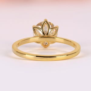 Könnte beinhalten: Ein goldener Ring mit blütenförmiger Fassung. Der Ring hat einen runden, hellfarbigen Edelstein, der von blütenblattartigen Zinken umgeben ist. Das Band ist aus einfachem, poliertem Gold. Der Ring ist vor einem weichen, verschwommenen Hintergrund platziert.