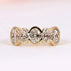 May include: A gold ring featuring five oval-cut diamonds. The diamonds are set in a row, with a polished gold band. The ring is displayed against a soft, light background, highlighting the sparkle of the gemstones.