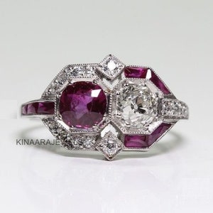 Peut inclure: Une bague de style vintage avec un grand diamant rond et deux rubis sertis dans une bande en platine. La bague est décorée de diamants et de rubis plus petits.