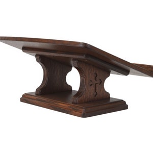 May include: A dark brown wooden lectern with a cross carved into the front of the base. The lectern has a slanted top surface for holding books or other materials.