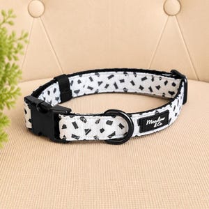 May include: A white pet collar featuring a black abstract geometric pattern. The collar includes a black plastic side-release buckle, a black metal D-ring, and a black fabric label with the white text Meadow & Co. stitched onto the strap.