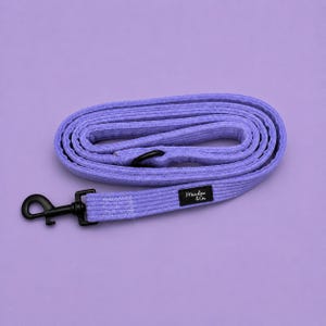 May include: A lavender corduroy dog lead coiled in a circular shape. The lead features a black metal swivel snap hook at one end and a black rectangular fabric label with the white text Meadow & Co. stitched onto the textured fabric strap.