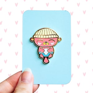 May include: A pink enamel pin of a cartoon otter wearing a yellow beanie and holding a white cloud. The otter has closed eyes and a pink nose.