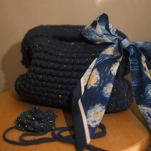 Può includere: Una borsa all'uncinetto blu navy scuro con sottili pagliuzze dorate nel filato. La borsa è decorata con un nastro di seta blu con un motivo vorticoso ispirato a una notte stellata annodato a fiocco e un piccolo fiore all'uncinetto abbinato.
