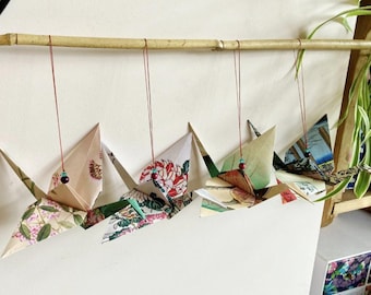 May include: Four origami cranes crafted from patterned paper, each suspended by a thin red string with a small bead. The cranes feature various floral and landscape designs in shades of pink, green, blue, and white, hanging from a bamboo rod.