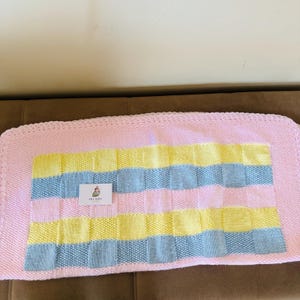 Peut inclure: Une couverture tricotée rectangulaire avec une bordure rose clair unie et un motif central en grille composé de carrés alternés jaune clair, bleu clair et rose clair. Une petite étiquette blanche avec le texte ERA BABY est fixée.