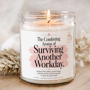 May include: A clear glass jar candle with a white wax fill and a lit wick. The label features black text reading The Comforting Aroma of Surviving Another Workday, a pink brushstroke graphic, a coffee mug icon, and additional descriptive text.