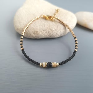 Peut inclure: Bracelet doré composé de petites perles noires et dorées. Il présente un motif central avec une perle noire encadrée de deux perles blanches. Le bracelet est posé sur un fond neutre.