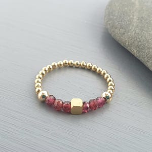 Peut inclure: Une bague dorée composée de perles et ornée d'une rangée centrale de pierres précieuses rose-rouge facettées et d'une perle carrée dorée. La bague est posée sur un fond gris, mettant en valeur le design et la couleur du bijou.