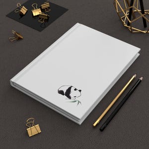 May include: A white notebook with a black and white illustration of a panda bear on the cover. The notebook is lying on a dark grey surface with two pencils and a gold metal decorative stand.