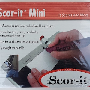 May include: A white and silver scoring tool with a red handle. The tool is labeled "Scor-it Mini" and "The Original Scor-it". The tool is used to score paper or cardstock for folding.