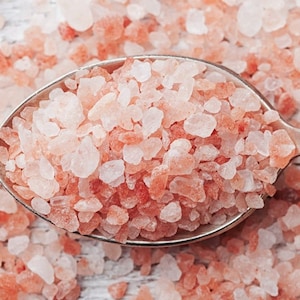 May include: A close-up of a silver spoon filled with pink Himalayan salt crystals. The salt is scattered around the spoon on a white wooden surface.