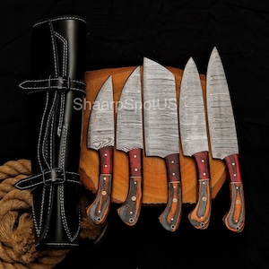 May include: A set of six Damascus steel kitchen knives with wooden handles and a black leather sheath. The blades have a unique pattern, and the handles are a mix of red and brown tones. The sheath is closed with a strap.