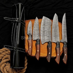 May include: A set of six Damascus steel kitchen knives with wooden handles, displayed on a wooden cutting board. The knives are accompanied by a black leather knife roll with white stitching. The text "SharpSpotUS" is visible.